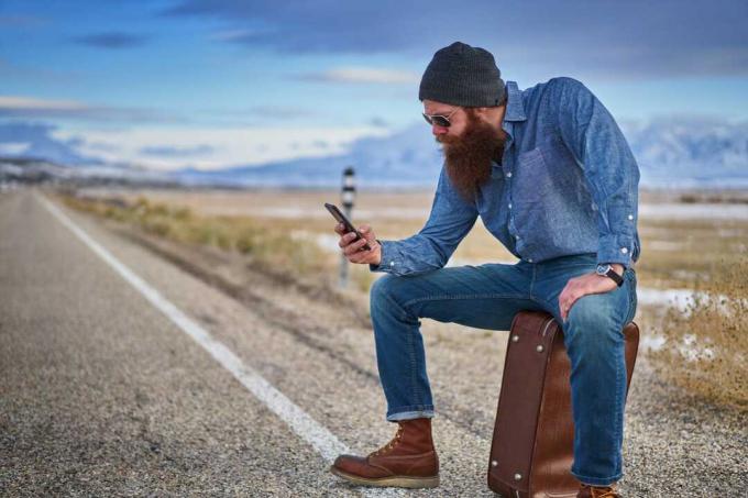 दाढ़ी वाला सहयात्री स्मार्ट फोन का उपयोग करते हुए सूटकेस पर बैठा है