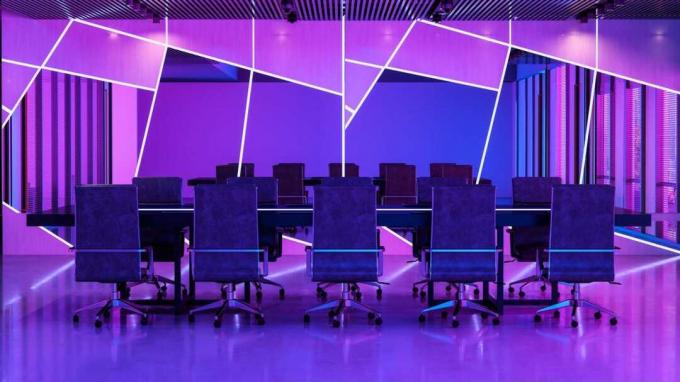 Eine Reihe von Schreibtischstühlen an einem Tisch in einem futuristischen, neonviolett beleuchteten Büro mit Spiegeln im Hintergrund, die in unregelmäßigen geometrischen Formen geschnitten sind